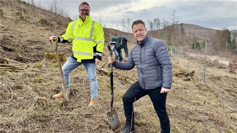 Zwei Männer mit Arbeitshandschuhen und Jacken stehen auf einem Hang, jeder hält eine Schaufel, im Hintergrund kahle Bäume und ein Zaun.