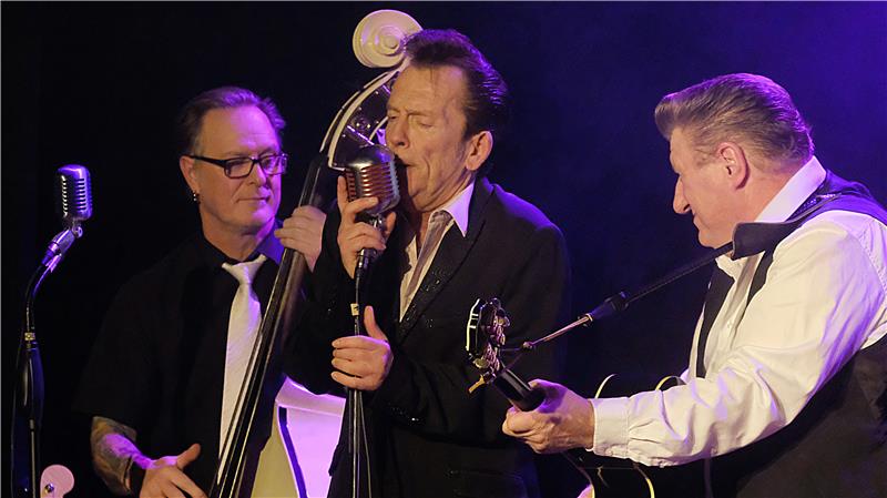 Drei Musiker stehen im violetten Lichtschein auf einer Bühne, sind in engem Kontakt und machen gemeinsam Musik mit Gesang und Saiteninstrumenten.