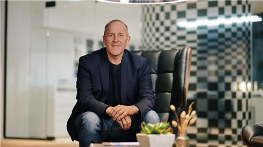 Mann sitzt mit verschränkten Händen auf einem Stuhl vor einem runden Tisch mit Pflanze und Dekoration in einem modernen Büro