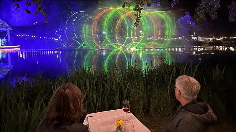 Das Foto zeigt ein Paar, das auf ein Gewässer mit Lasershow schaut. 