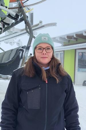 Person mit hellgrüner Mütze und dunkler Jacke steht vor einem Sessellift in einer verschneiten Winterlandschaft.