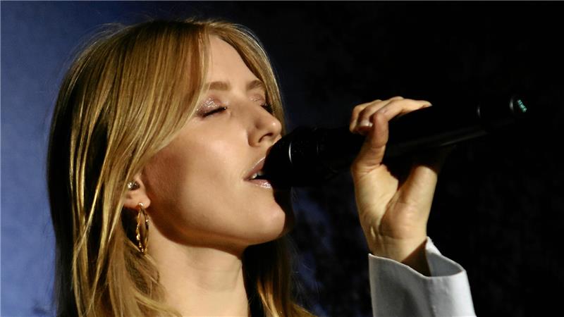 Frau mit langen blonden Haaren trägt eine helle Jacke und hält ein Mikrofon in der linken Hand