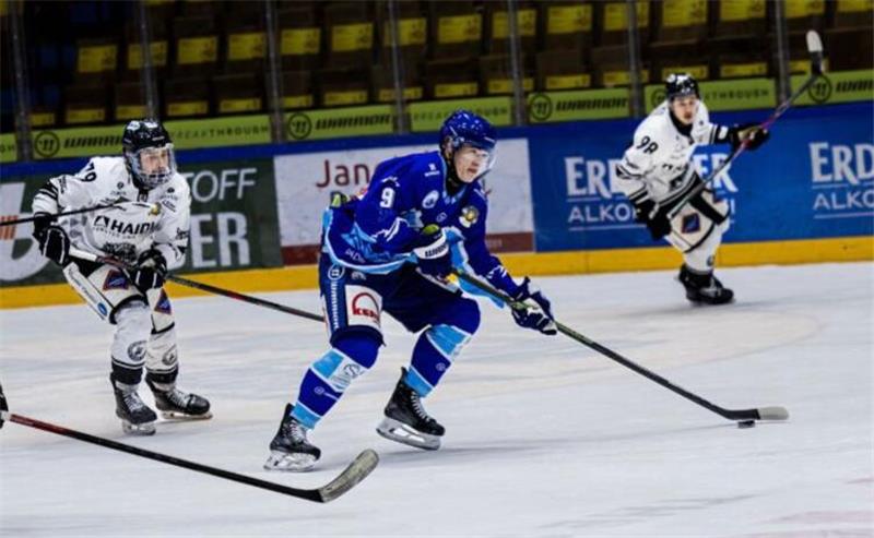 Eishockeyspieler in blauen und weißen Trikots kämpfen um den Puck auf dem Spielfeld in einer Halle mit leeren Zuschauerrängen.