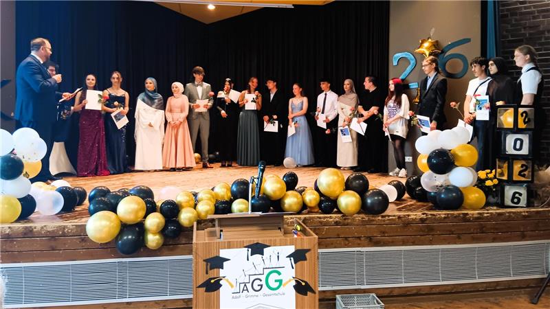 Gruppe von jungen Menschen in festlicher Kleidung steht auf einer Bühne mit Ballon-Dekoration und einem Redner am linken Rand