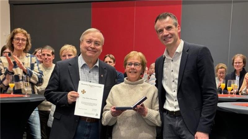 Hartes Training für Schwimmer und große Ehre für Schwarzmann Drei Personen stehen in einem Raum mit grauem Teppich und roter Wand, die mittlere Person hält eine Urkunde und eine Schachtel, im Hintergrund stehen weitere Personen an Stehtischen mit schwarzen Hussen.