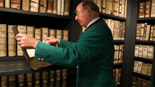 Älterer Mann mit Brille und grünem Sakko zieht ein großes Buch aus einem Regal voller alter Bücher.