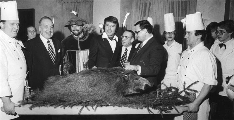 Gruppe von Personen in formeller Kleidung und Kochuniformen trägt einen großen Wildschweinkörper auf einem Tablett mit Tannenzweigen