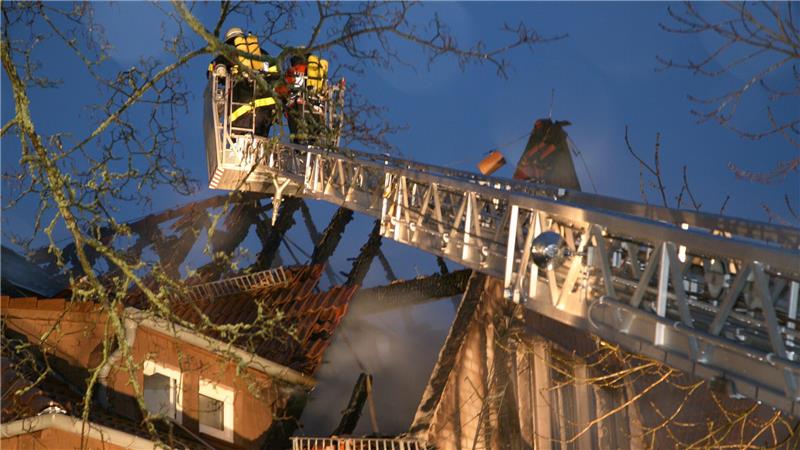 Feuerwehrleute auf einer ausgefahrenen Drehleiter über einem brennenden Hausdach in der Dämmerung