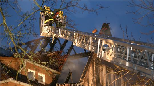 Feuerwehrleute auf einer ausgefahrenen Drehleiter über einem brennenden Hausdach in der Dämmerung