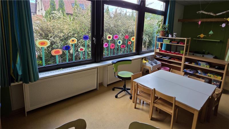 Kindgerechter Raum mit mehreren bunten Blumenaufklebern an großen Fenstern, kleinen Holztischen und Stühlen, einem grünen Stuhl auf Rollen und einem Regal mit Spielzeug.