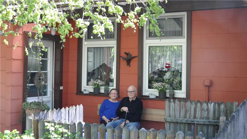 Älteres Paar sitzt auf einer Bank vor einem rotbraunen Haus mit zwei Fenstern und einem kleinen Holzzaun im Vordergrund