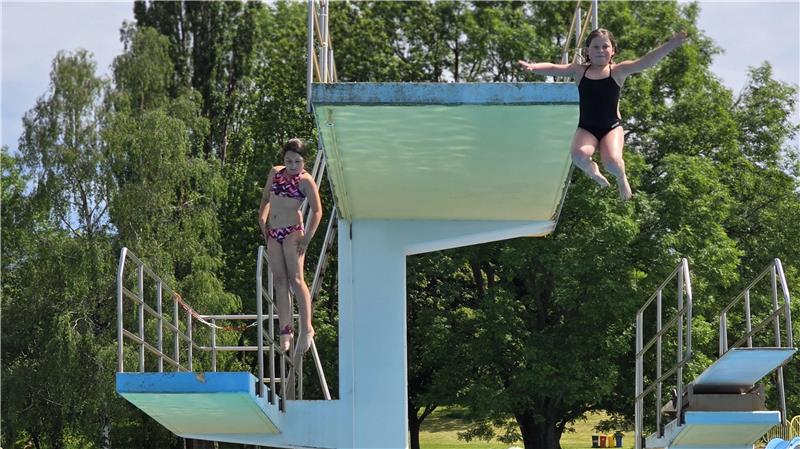 Zwei Mädchen springen von verschiedenen Sprungbrettern eines Schwimmbeckens, umgeben von Bäumen und Wiese.
