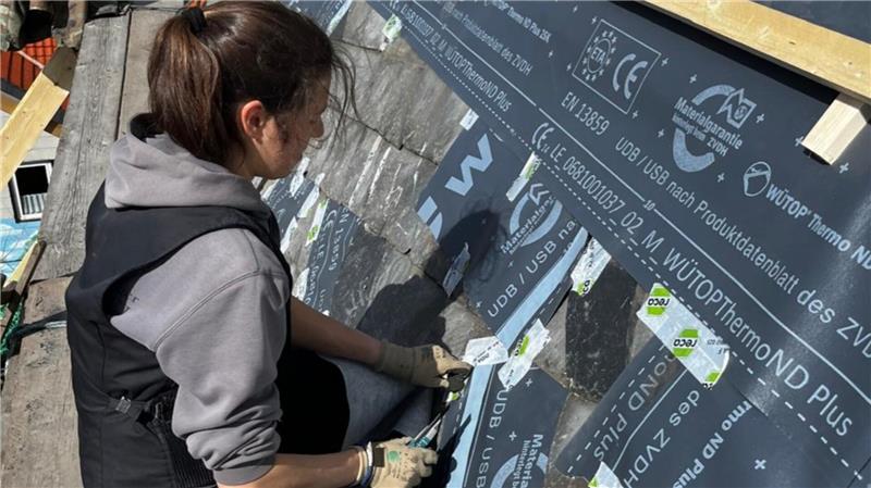 Frau mit Zopf und grauer Kapuzenjacke arbeitet auf einem Dach mit schwarzer Dämmfolie, die mit weißem Text bedruckt ist.