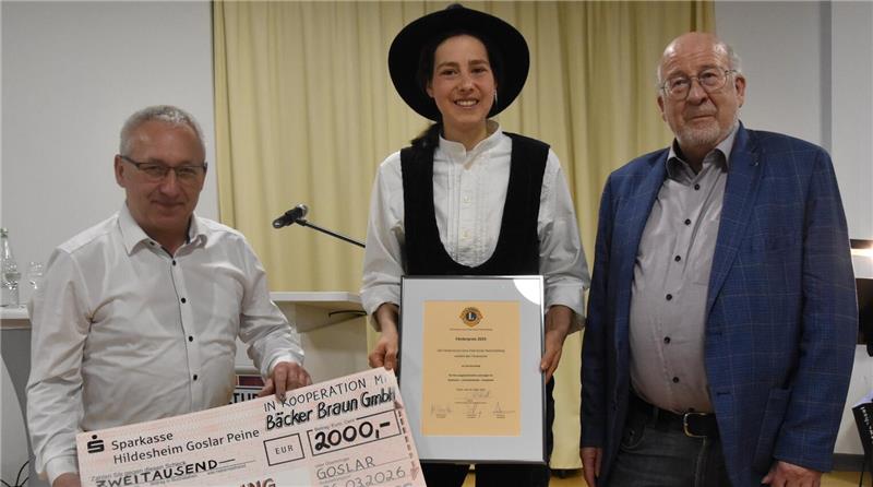 Person in traditioneller Tracht hält eine Urkunde, flankiert von zwei Personen in moderner Kleidung, eine davon hält einen großen Scheck mit der Aufschrift 'ZWEITAUSEND'.