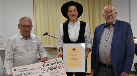 Person in traditioneller Tracht hält eine Urkunde, flankiert von zwei Personen in moderner Kleidung, eine davon hält einen großen Scheck mit der Aufschrift 'ZWEITAUSEND'.