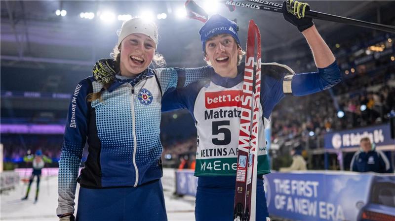 Zwei Skilangläufer in Wettkampfkleidung stehen nebeneinander in einer Halle, einer hält einen Skistock und Skier, im Hintergrund Zuschauertribüne und Werbebanner
