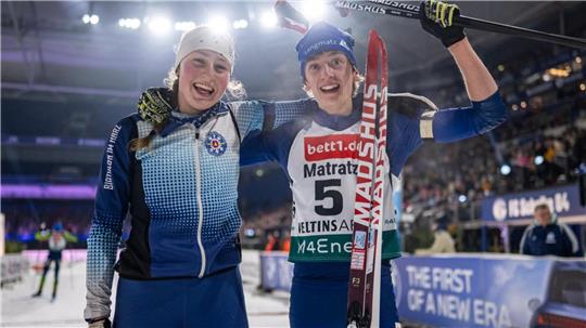 Zwei Skilangläufer in Wettkampfkleidung stehen nebeneinander in einer Halle, einer hält einen Skistock und Skier, im Hintergrund Zuschauertribüne und Werbebanner
