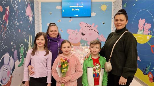 Fünf Personen stehen vor einer Wand mit Peppa-Wutz-Motiven und einem Bildschirm mit der Aufschrift 'HAPPY BIRTHDAY Olivia'.