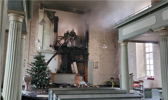 Löscharbeiten in der Beuchter Kirche an Weihnachten 2024, neben dem verkohlten Altar steht der Weihnachtsbaum, Rauch zieht durch das Kirchenschiff.