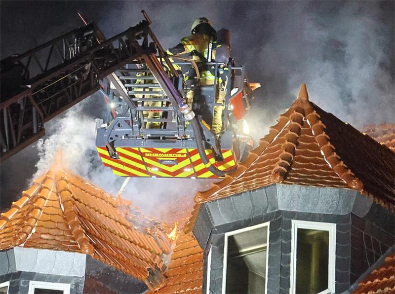 Feuerwehrleute in einem Hubsteiger an einer Drehleiter über einem Dach mit Rauchentwicklung bei Dunkelheit.