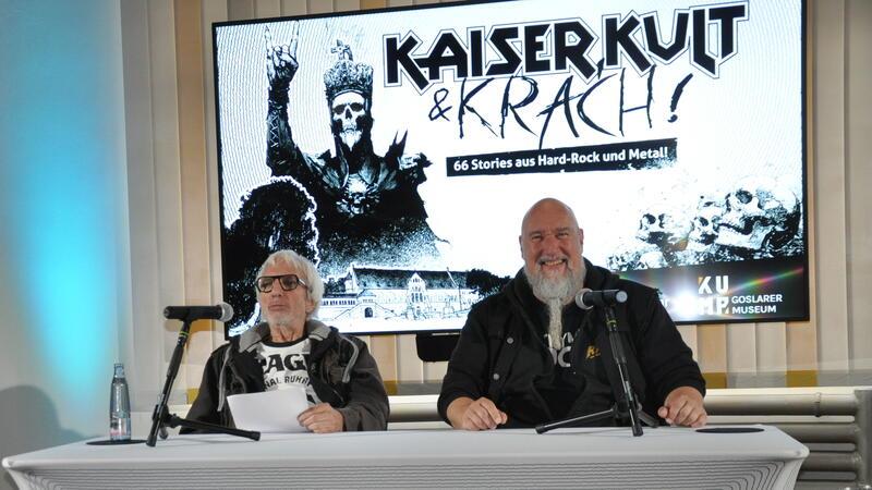 Zwei Personen sitzen an einem Tisch mit Mikrofonen vor einem Bildschirm mit der Aufschrift 'KAISER KULT & KRACH! 66 Stories aus Hard-Rock und Metal'