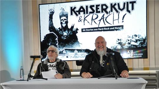 Zwei Personen sitzen an einem Tisch mit Mikrofonen vor einem Bildschirm mit der Aufschrift 'KAISER KULT & KRACH! 66 Stories aus Hard-Rock und Metal'
