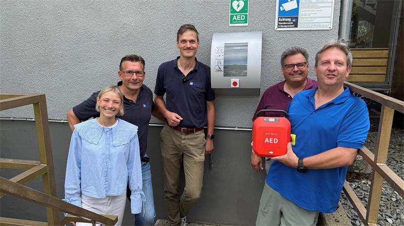 Das Foto zeigt eine Frau und vier Männer, von denen einer einen Defibrillator in der Hand hält.
