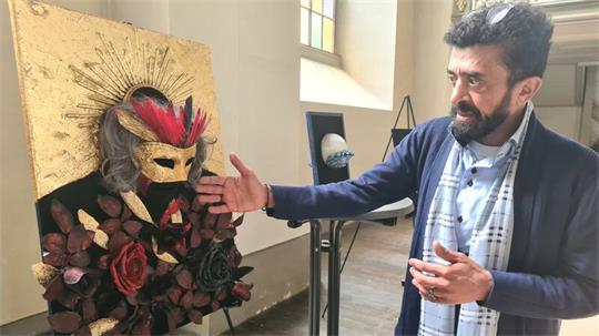 Ein Mann im Hemd und blauem Jacket erläutert ein Kunstwerk mit einer goldenen Maske, Federn und Rosen.