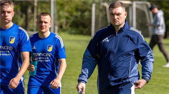 Zwei Personen in blauen Sporttrikots und eine Person in blauem Trainingsanzug auf einem Fußballfeld.