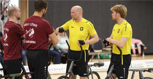 Vier Radballspieler in Sportkleidung stehen mit ihren Fahrrädern in einer Halle und unterhalten sich.