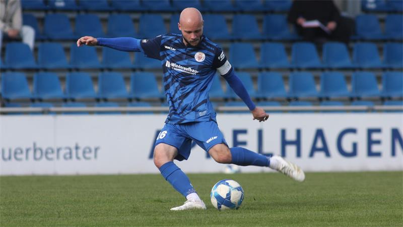 Fußballspieler in blauem Trikot mit Kapitänsbinde schießt Ball auf dem Spielfeld.