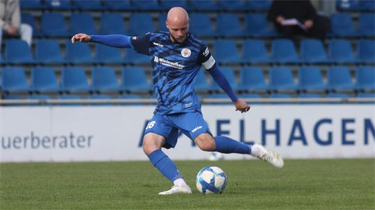 Fußballspieler in blauem Trikot mit Kapitänsbinde schießt Ball auf dem Spielfeld.