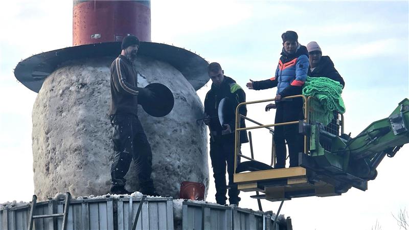 Vier Personen stehen auf einer Plattform vor einem großen, runden, steinernen Sockel mit einem zylindrischen Turm darauf, eine Person hält eine schwarze Scheibe.