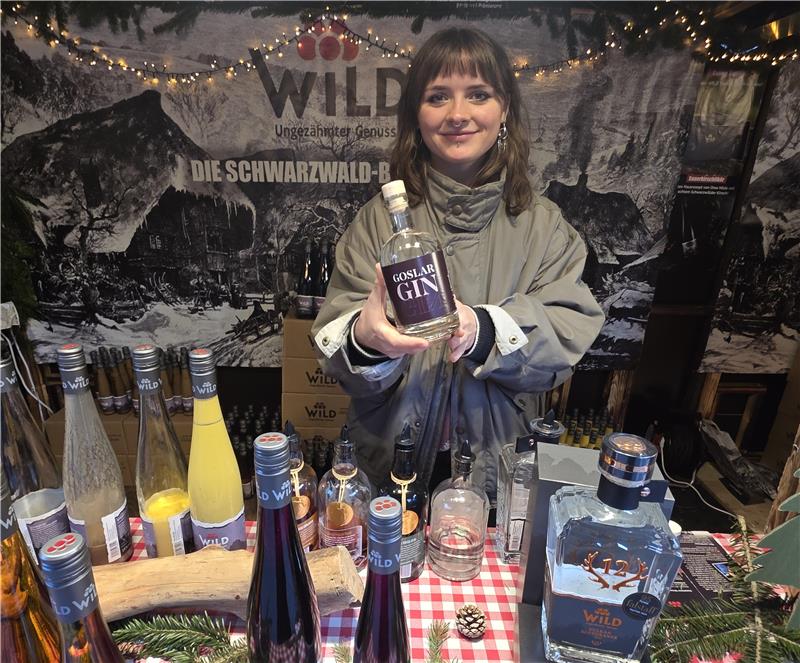 Person hält eine Flasche Gaslan Gin vor einem Stand mit verschiedenen Spirituosenflaschen und einem schwarz-weißen Schwarzwald-Banner im Hintergrund.