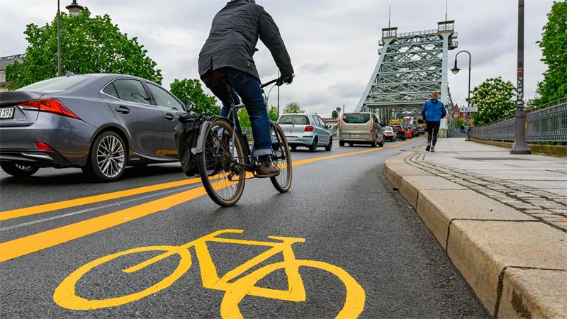 Fahrradfahrer auf einem Radweg mit gelbem Fahrradsymbol auf der Straße neben Autos und einer Brücke im Hintergrund