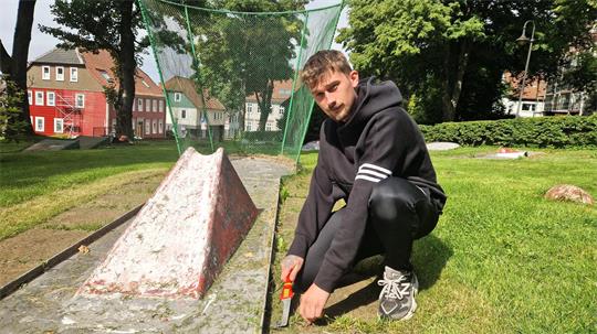 Ein junger Mann kniet neben einer gammeligen Minigolfbahn mit einem Gartenwerkzeug in der Hand.