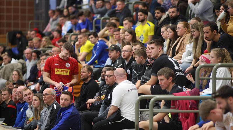 Zuschauer sitzen auf Tribünen in einer Sporthalle, einige tragen Sportkleidung in Rot, Gelb und Blau.