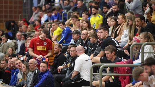 Zuschauer sitzen auf Tribünen in einer Sporthalle, einige tragen Sportkleidung in Rot, Gelb und Blau.