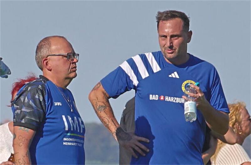 Zwei Männer in blauen Sporttrikots stehen an einer Absperrung, einer trinkt aus einer Flasche, im Hintergrund stehen weitere Zuschauer.