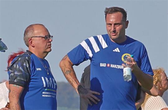 Zwei Männer in blauen Sporttrikots stehen an einer Absperrung, einer trinkt aus einer Flasche, im Hintergrund stehen weitere Zuschauer.