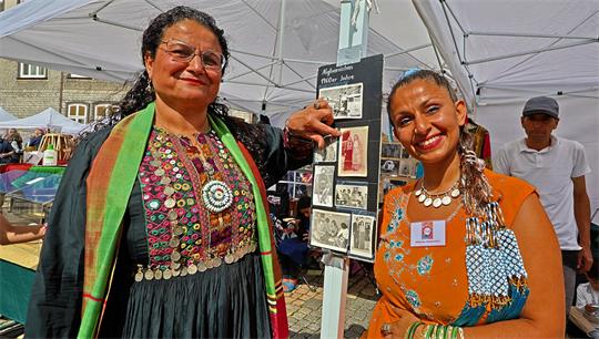 Zwei Frauen in farbenfroher traditioneller Kleidung stehen vor einem Marktstand mit Postkarten und lächeln in die Kamera
