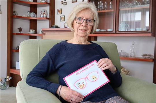 Ältere Person sitzt auf einem grünen Sofa und hält ein Buch mit einer Illustration von einer Brille und dem Text 'Die rosarote Brille'