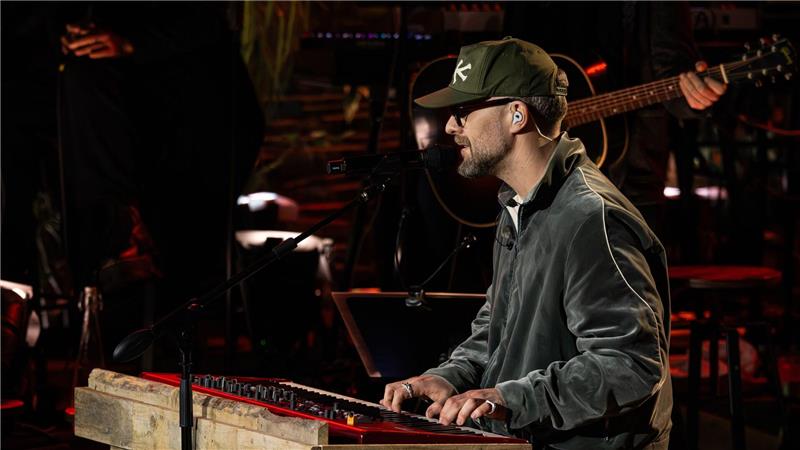 Musiker mit Baseballkappe spielt Keyboard auf einer Bühne mit dunklem Hintergrund.