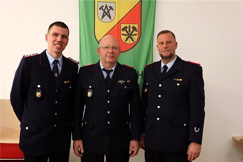 Drei Feuerwehrleute vor Ortswappen. 