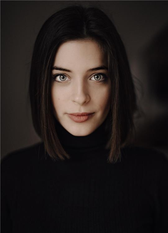 Weibliche Person mit schulterlangem, glattem Haar trägt schwarzen Rollkragenpullover vor dunklem Hintergrund.