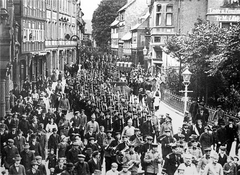 Große Menschenmenge mit uniformierten Soldaten in einer historischen Straßenparade zwischen mehrstöckigen Gebäuden