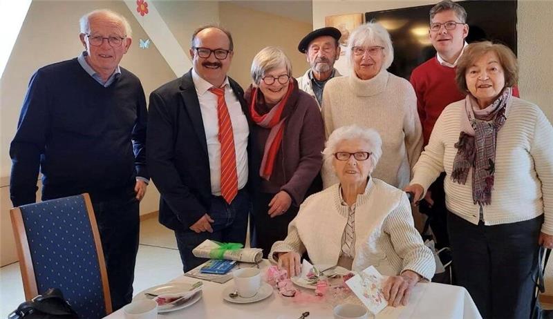 Gruppe von acht Personen, davon eine ältere sitzend am gedeckten Tisch, die anderen stehend, in einem Raum mit schräger Decke