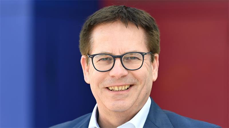 Ein lächelnder Mann mit Brille trägt ein weißes Hemd und einen blauen Anzug vor einem unscharfen Hintergrund in Blau und Rot