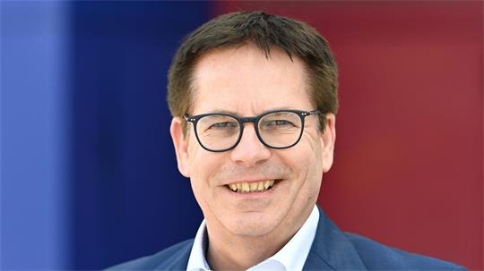 Ein lächelnder Mann mit Brille trägt ein weißes Hemd und einen blauen Anzug vor einem unscharfen Hintergrund in Blau und Rot