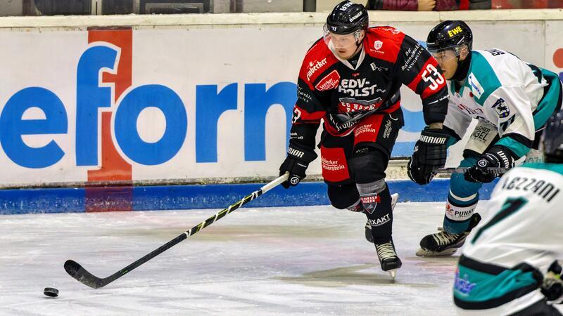 Eishockeyspieler in schwarzer und roter Ausrüstung mit Schläger vor Puck auf Eisfläche, zwei Gegner in weißer und grüner Ausrüstung verfolgen ihn